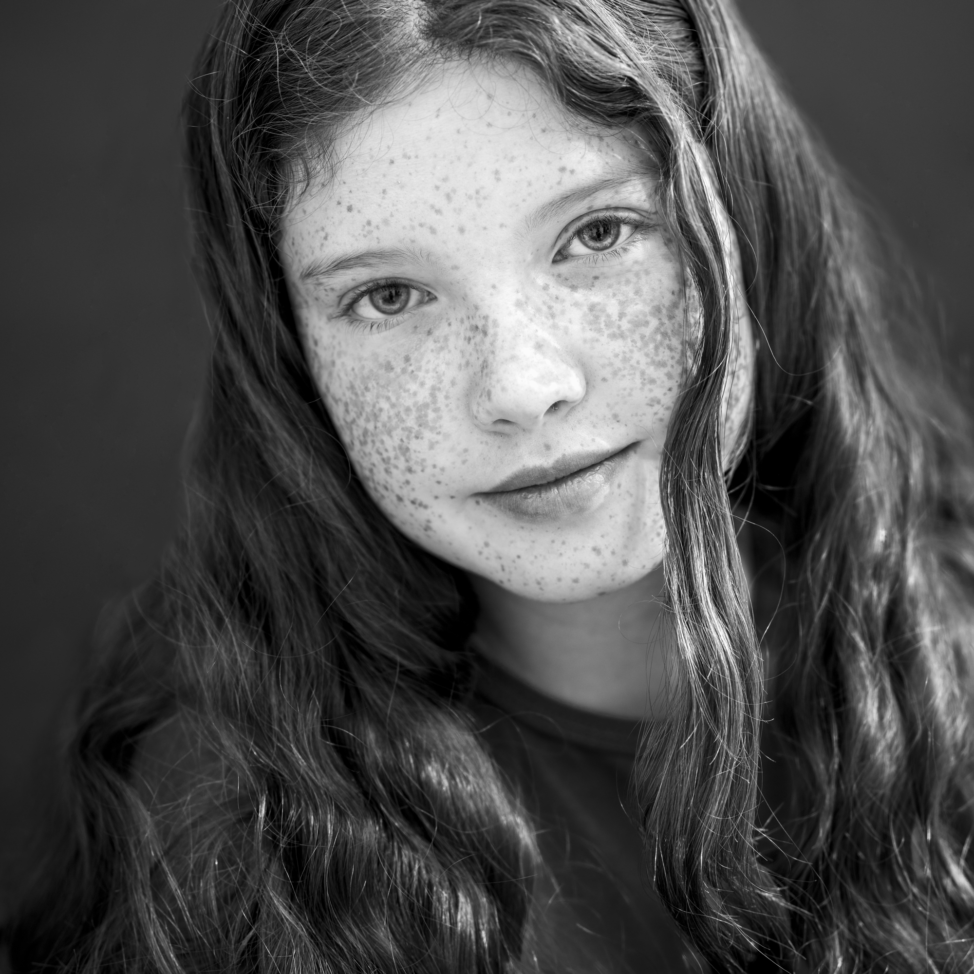 Black-and-white portrait of a young girl with long wavy hair and freckles, looking softly at the camera, by Jerome Art Photography.