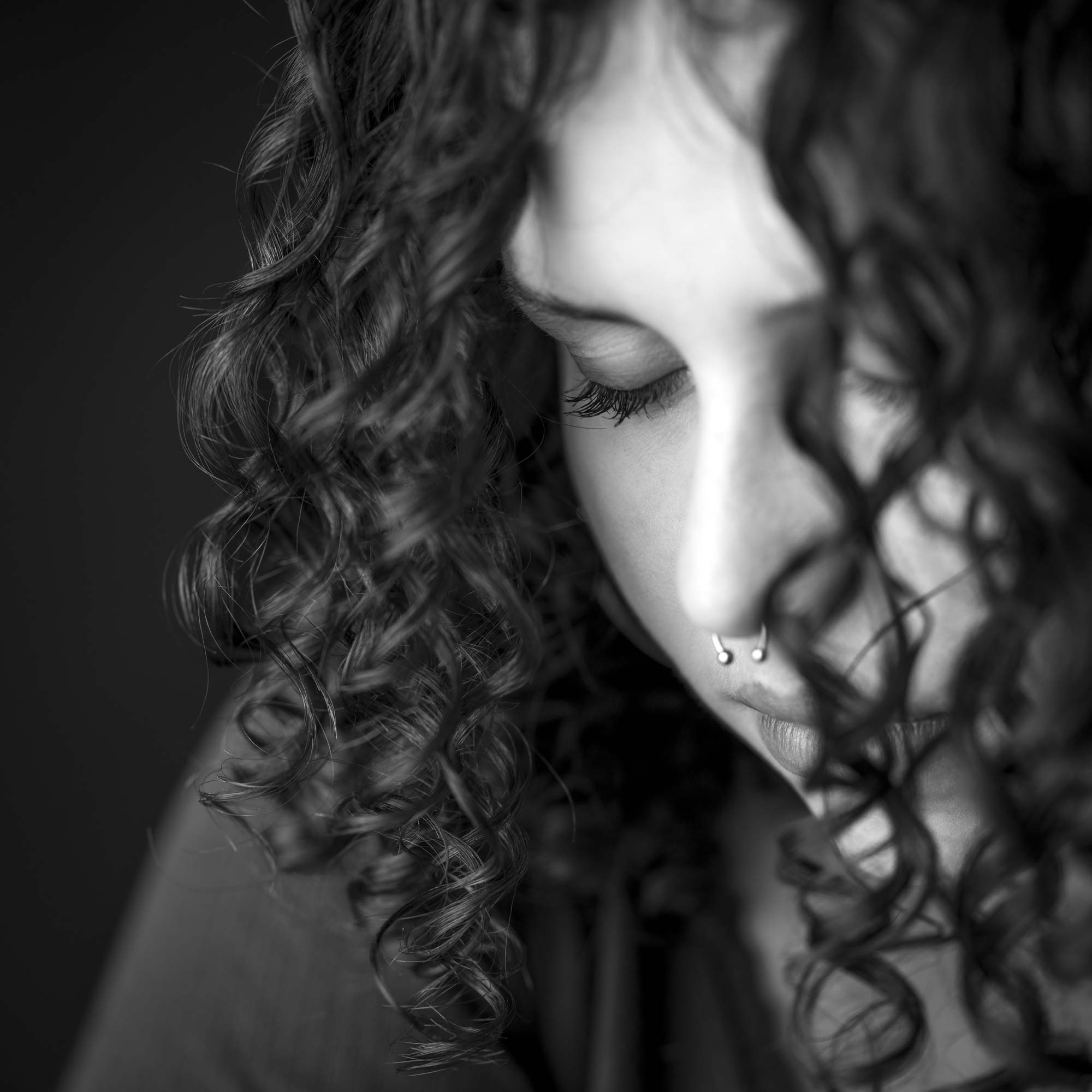 Black-and-white close-up portrait of a young woman with curly hair and a nose piercing, eyes closed in a contemplative expression, by Jerome Art Photography.