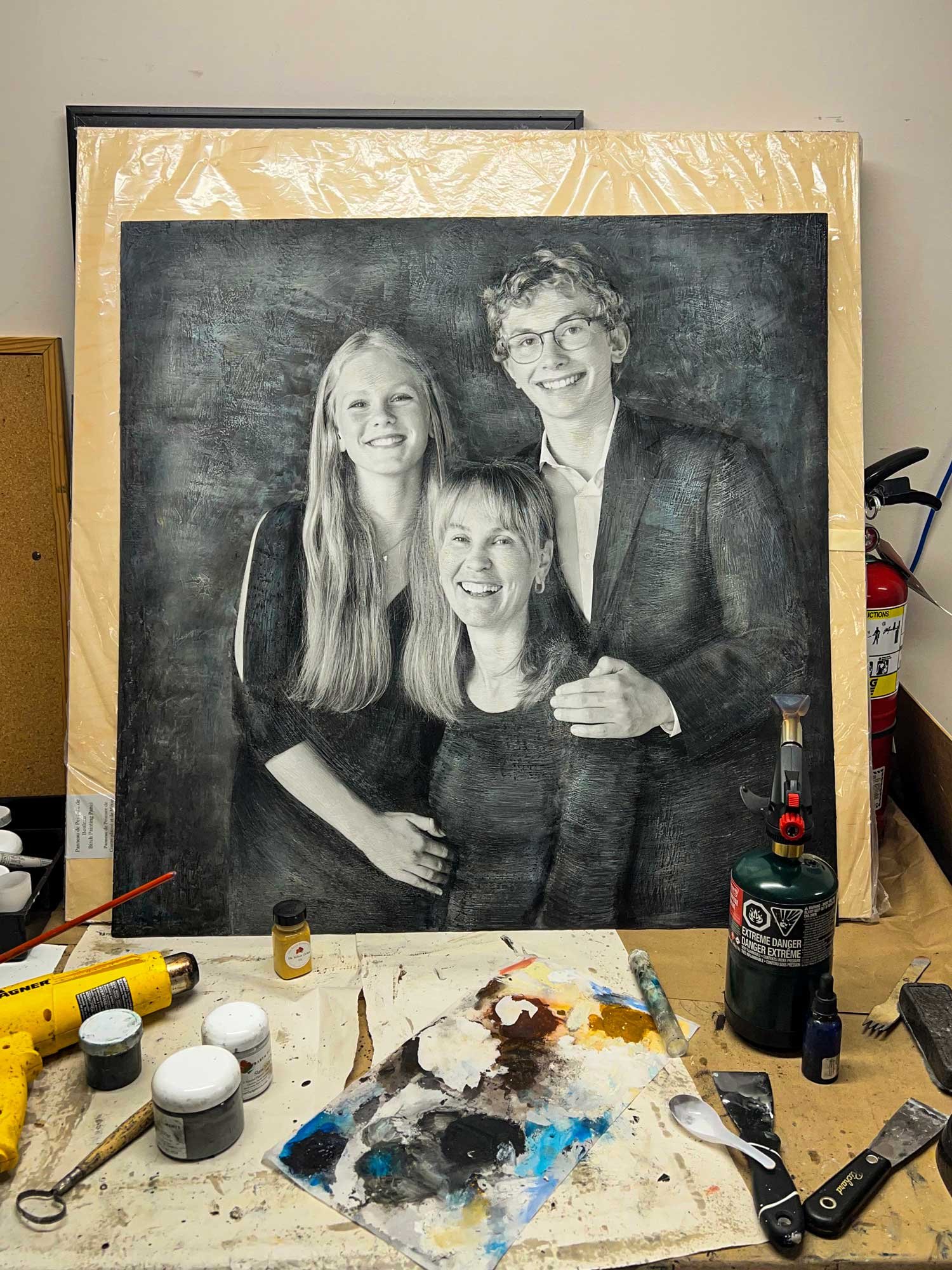 Artist’s workspace featuring a large mixed media family portrait in black and white, surrounded by paint supplies, brushes, and tools.