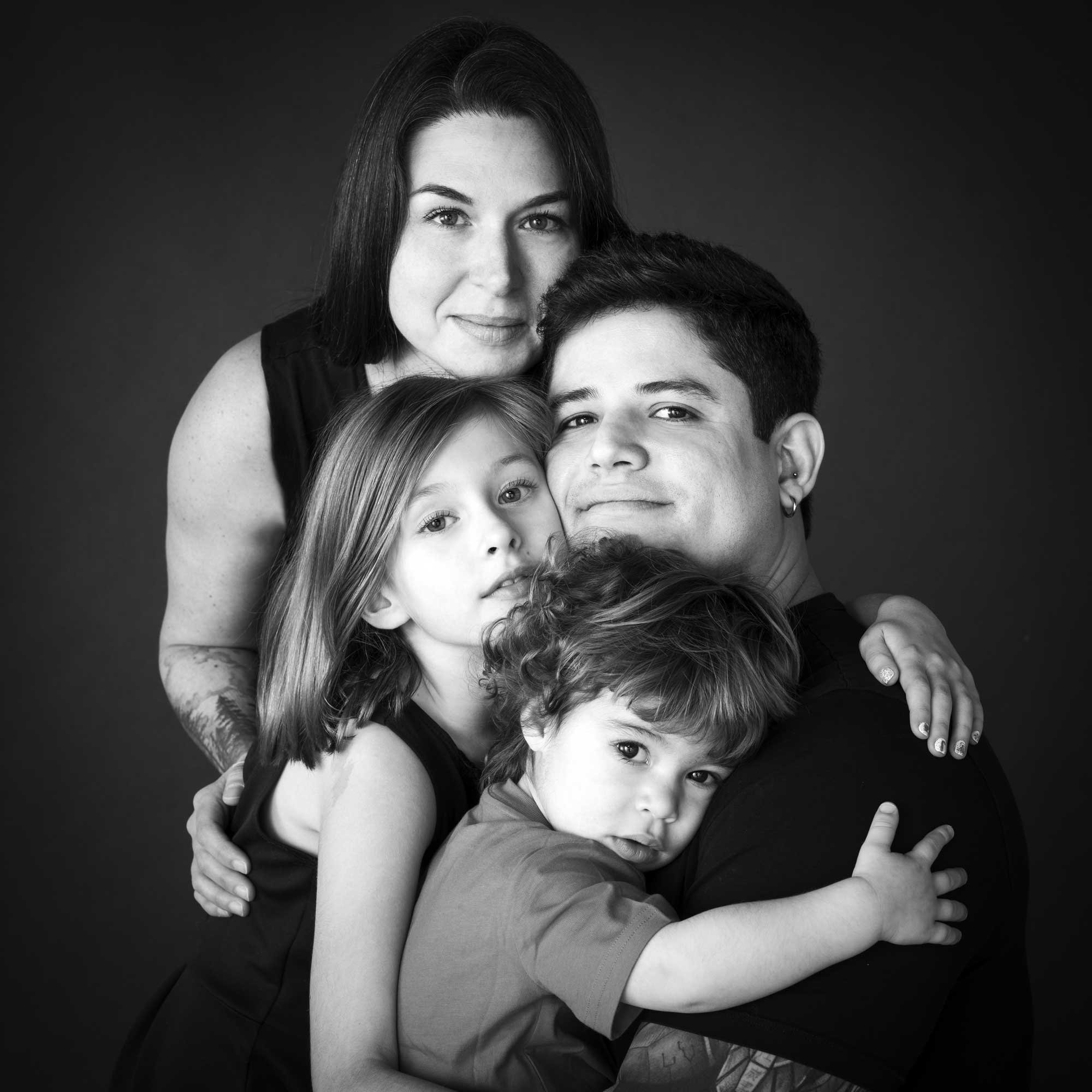 Black-and-white portrait of a family of four embracing each other, with the mother in the background and the father holding two children closely, by Jerome Art Photography.