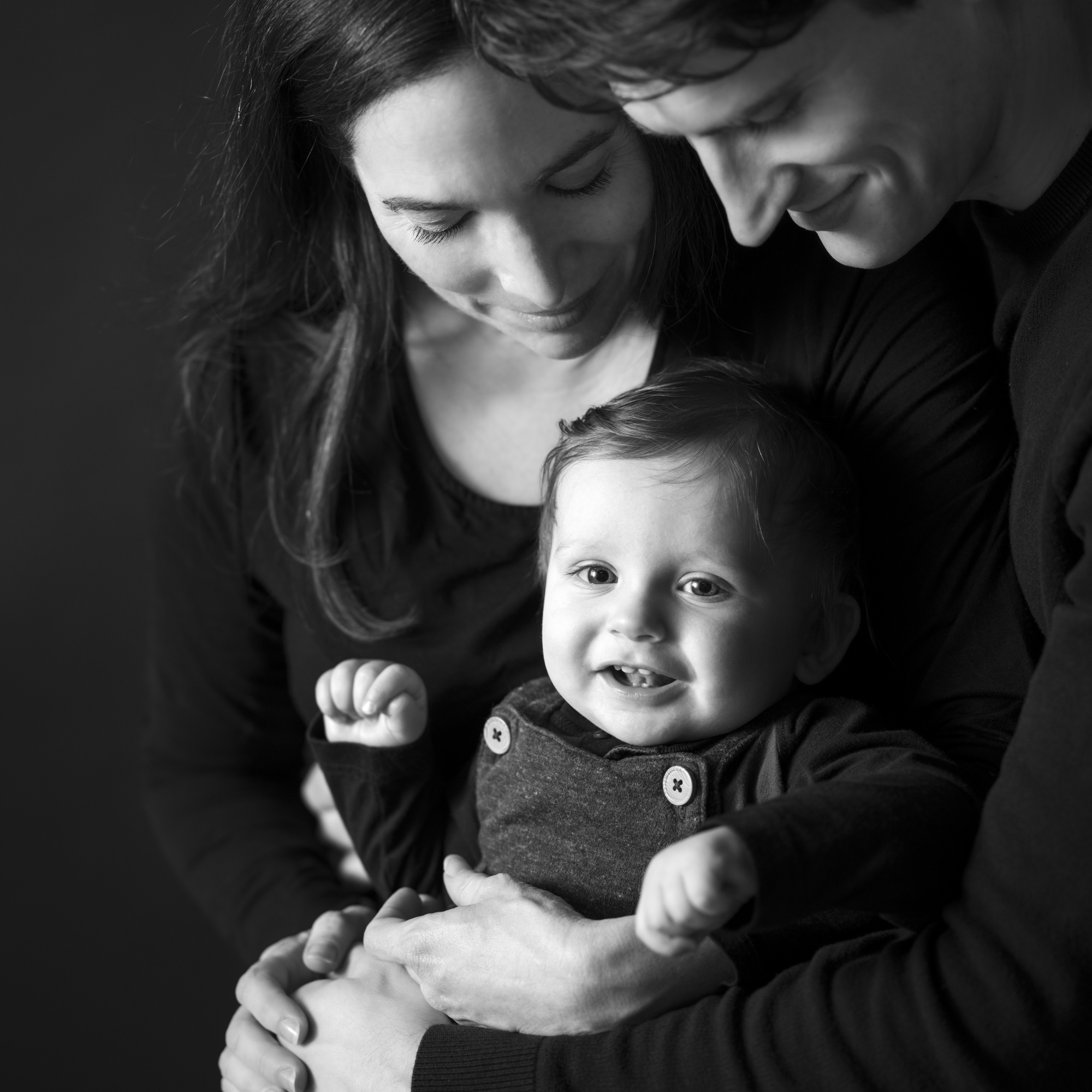 Black-and-white portrait of a smiling baby held in the arms of loving parents, all wearing dark clothing, by Jerome Art Photography.