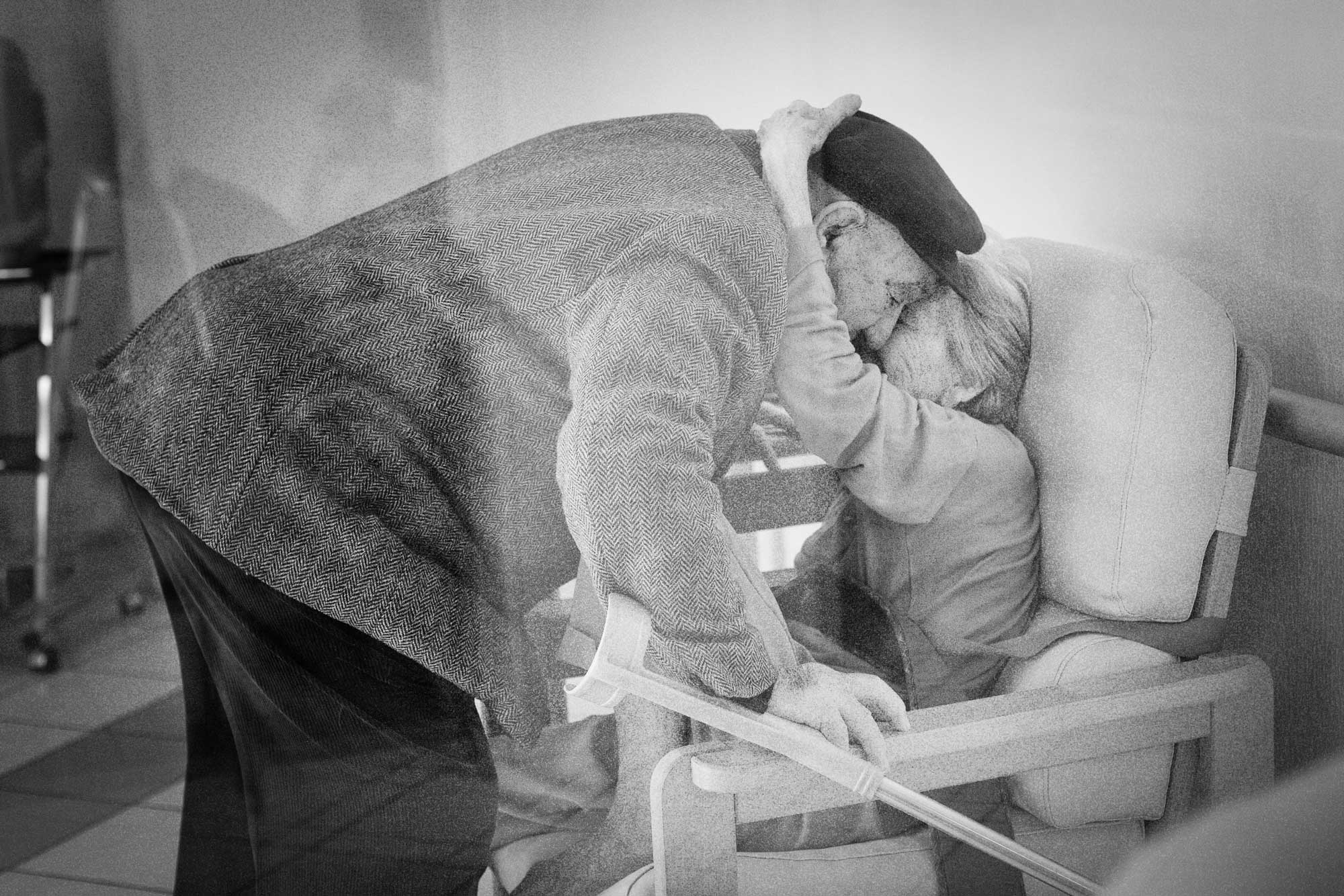 75 Years of Love – Intimate Black & White Portrait by Jérôme Elderly man and woman embracing tenderly through a care home window, symbolizing 75 years of enduring love