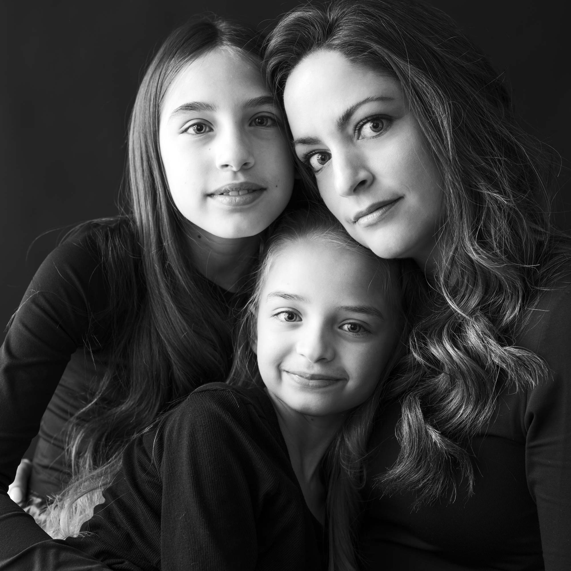 Portrait en noir et blanc d'une mère et de ses deux filles, toutes vêtues de noir, étroitement enlacées, par Jerome Art Photography.