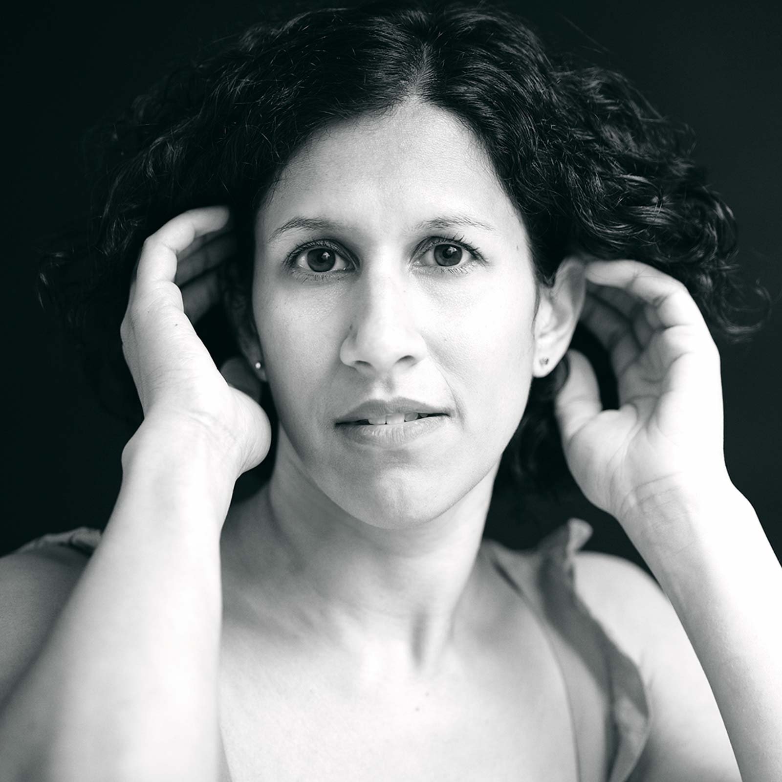 Portrait en noir et blanc d'une femme aux cheveux courts et bouclés, regardant directement l'appareil photo, les mains levées à côté de son visage.