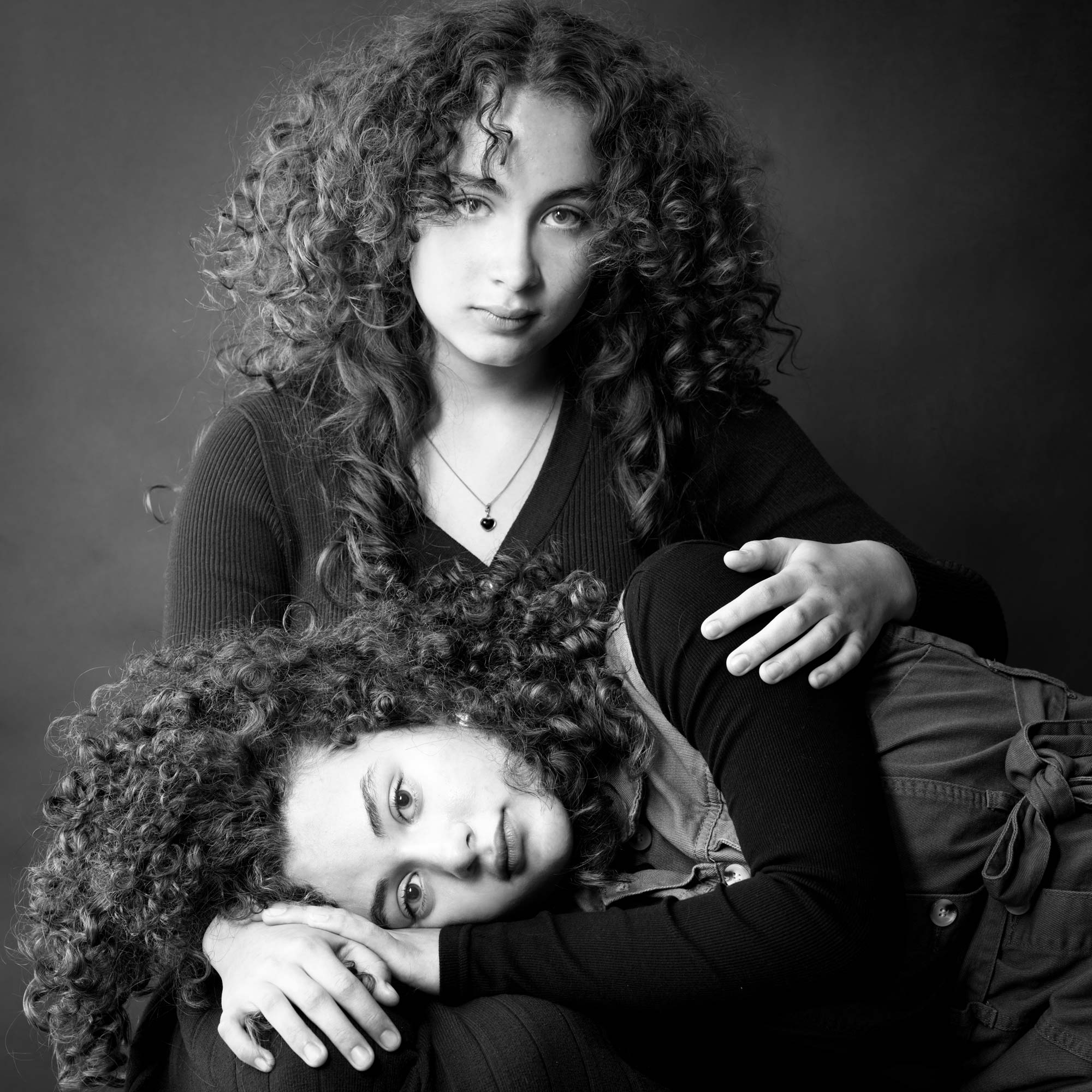Portrait en noir et blanc de deux jeunes filles aux cheveux bouclés, l'une assise et l'autre reposant sa tête sur les genoux de sa sœur, par Jerome Art Photography.