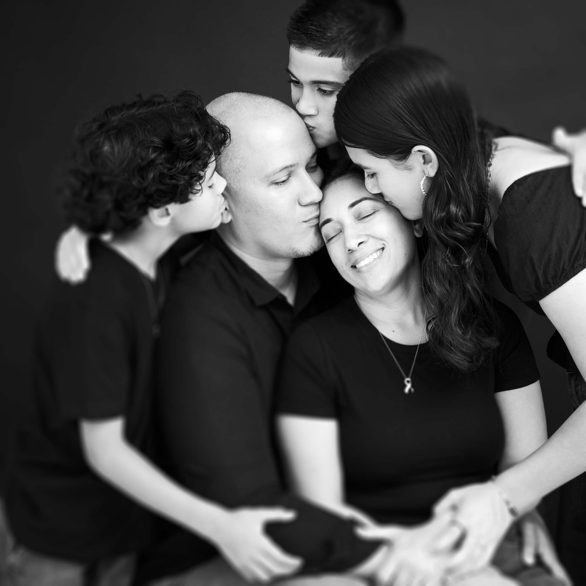 Black-and-white family portrait: parents seated, children leaning in to hug and kiss, eyes closed in a calm embrace.