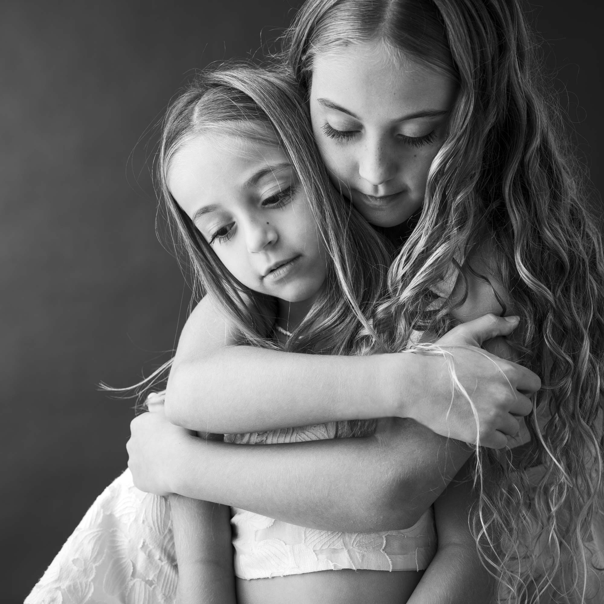 Two young sisters with long hair share a tender embrace in black and white, expressing love, connection, and quiet emotion.