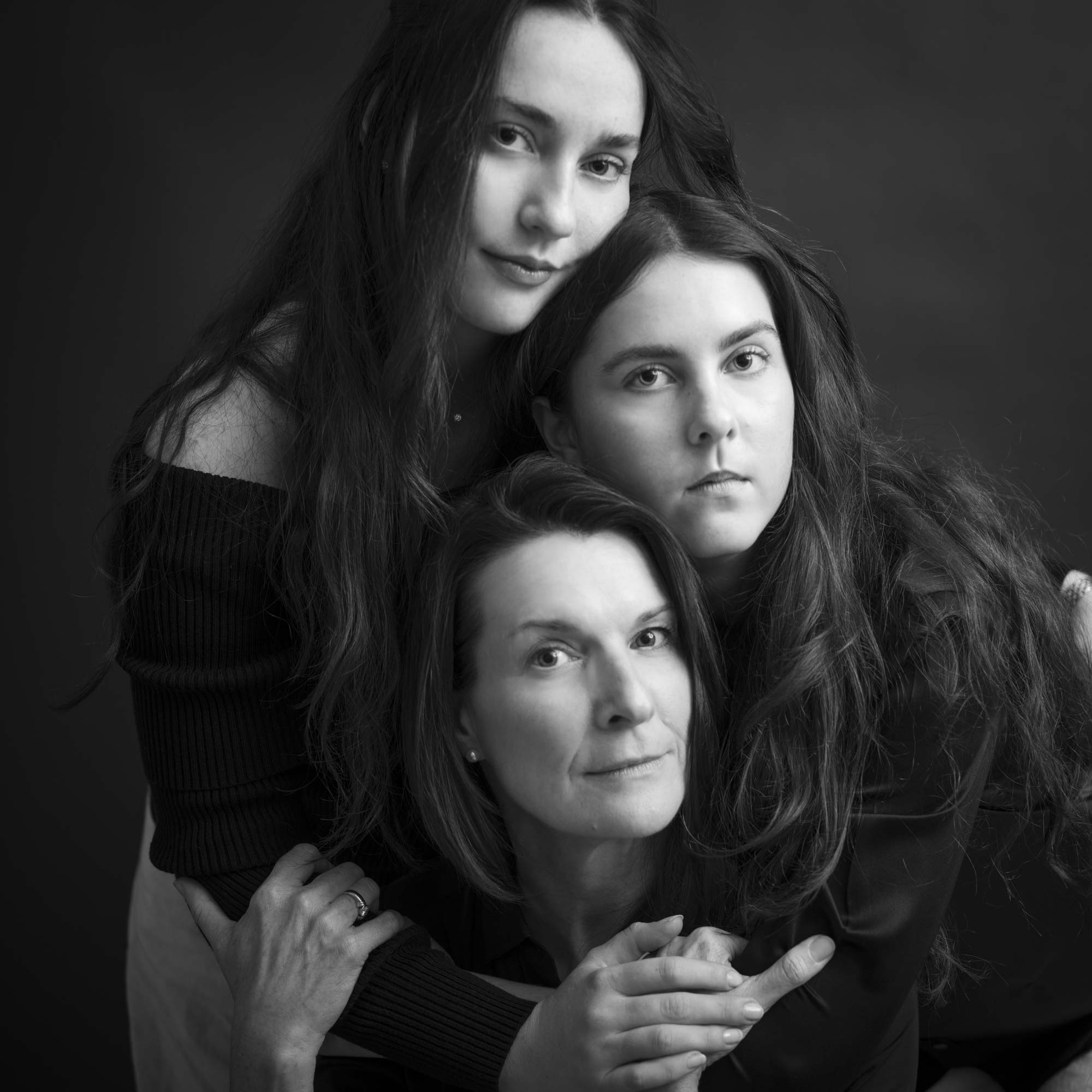 Fine art black and white studio portrait of a mother embraced by her two daughters, all looking toward the camera with calm, intimate expressions