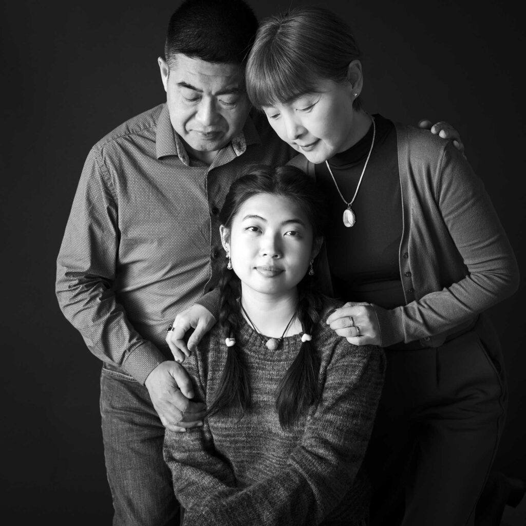 Black and white studio portrait of an adult daughter seated in front, looking at the camera, while her mother and father stand behind her, leaning in and looking down toward her with gentle expressions.