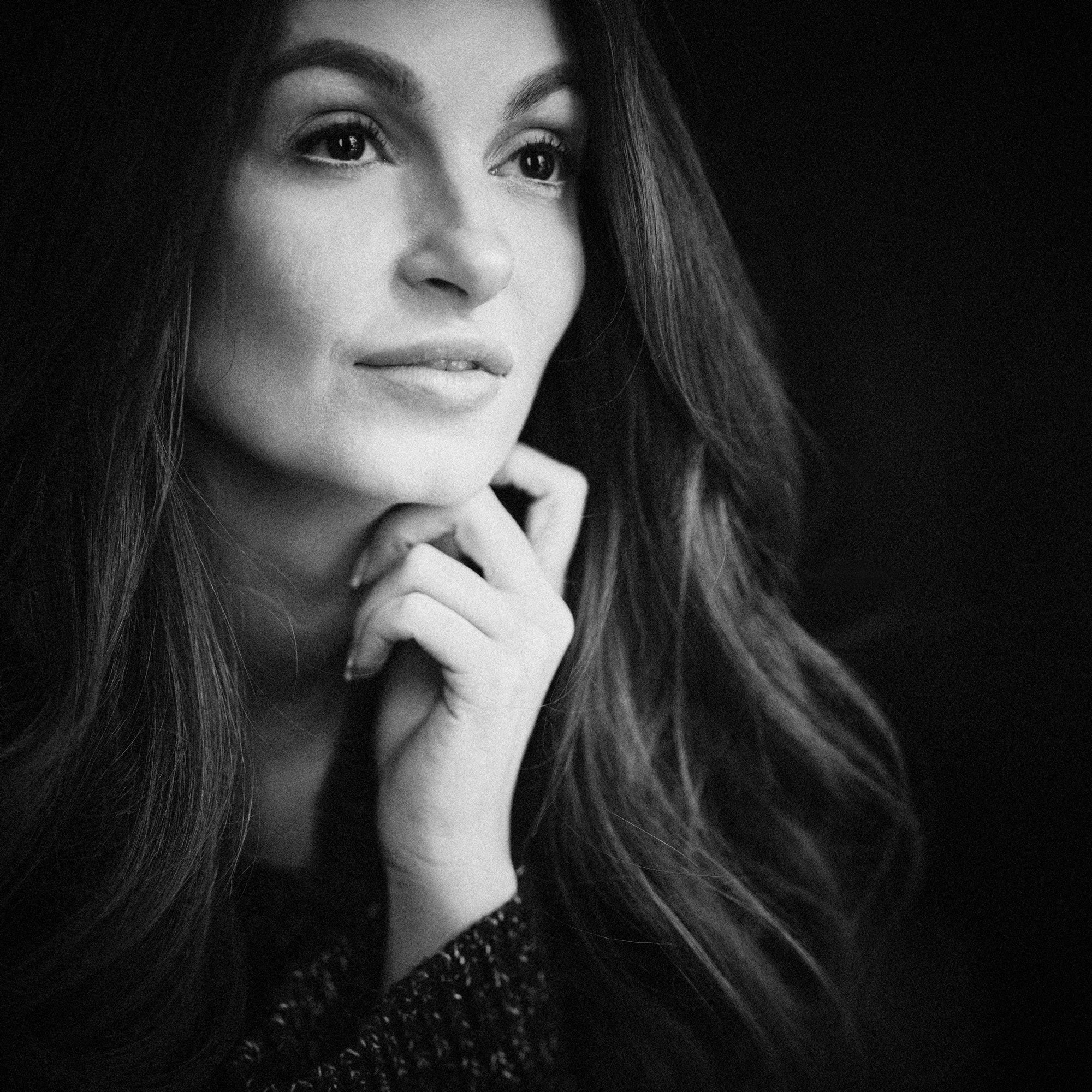 Soft grain black and white close up portrait of a woman with long hair, resting her chin on her hand and gazing toward the light, created as an intimate beauty portrait by Jérôme Scullino.
